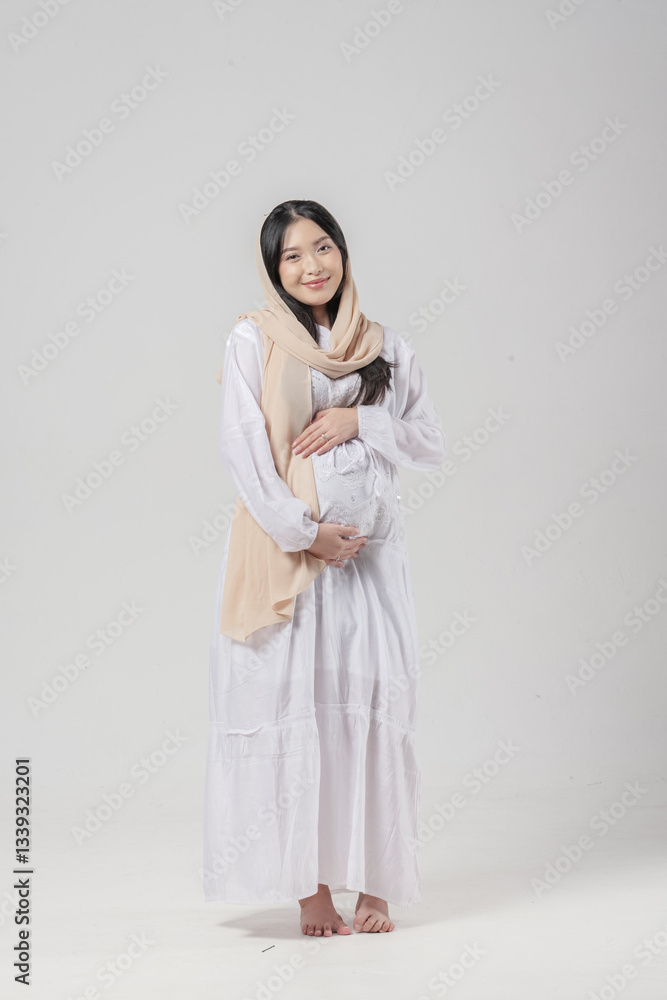 Happy Asian Muslim pregnant woman wearing veil hijab smiling while touching her stomach.