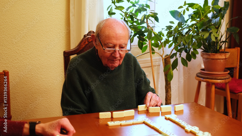Concentrated elderly man arranging domino tiles on a wooden table at home, enjoying his leisure time