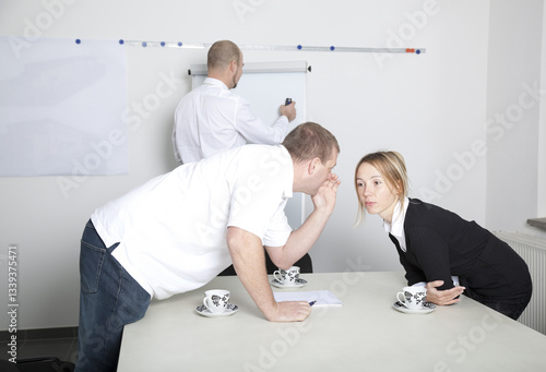 Secret conversation in the workplace. Two colleagues whisper at a table during a work meeting while a third person writes on a board. 