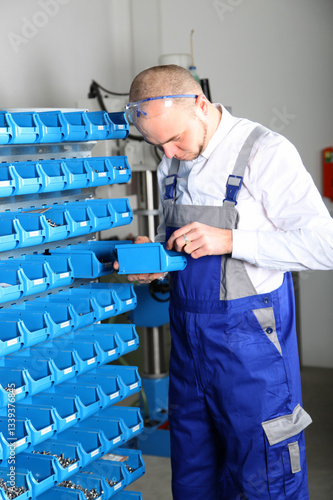 Worker sorting parts in storage bins. A man in work clothes stands by a shelving unit with plastic storage bins for small parts. He is carefully searching for or placing an item in one of the bins.