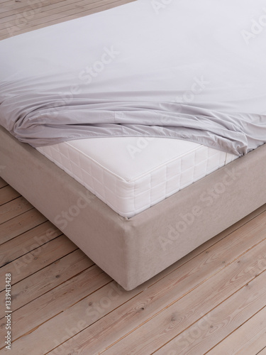 Bed in a bright bedroom with wooden floors is covered with grey fitted sheet with an elastic band, but the corner of the white mattress is visible. Side view photo. Suitable for mock-up.