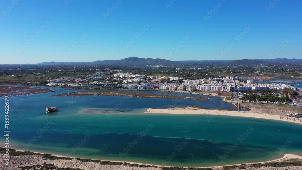Aerial view of Fuseta Portugal showing the coastal town with white buildings a sandy beach turquoise waters a historic structure in the lagoon a marina and surrounding green hills Aerial view of Fuseta Portugal showing the coastal town with white buildings a sandy beach turquoise waters a historic structure in the lagoon a marina and surrounding green hills