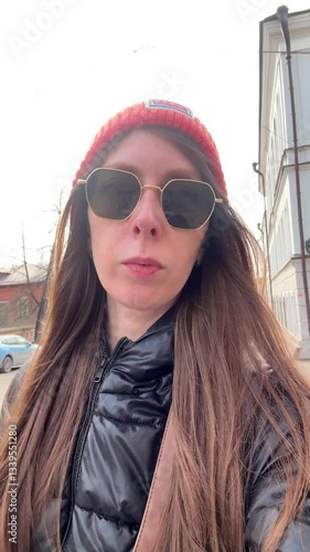young and beautiful woman in sunglasses, orange hat and black jacket, walks around the city and makes video selfies. Travels of a lonely girl around the cities