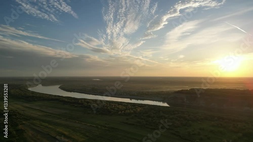 The sun sets over a meandering river, casting warm golden light on the surrounding fields and distant hills