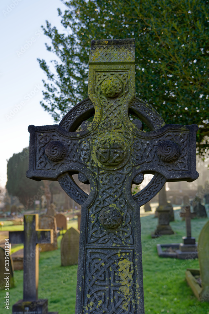Old stone cross at church graveyard in Swindon, UK. Cross as religious symbol of Christianity. Sun rays on cemetery in the background on a sunny morning. Beautiful stonework.