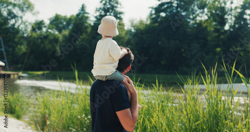 Photography Caring father carries his son on his shoulder, wearing a hat, by a pond watching surfing competitions in the midst of nature