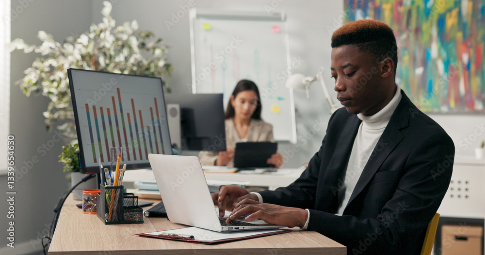 A smiling dark-skinned boy spends morning at work in front of computer screen, tapping fingers on laptop keyboard, filling out official and company applications, comparing correctness on two monitors A smiling dark-skinned boy spends morning at work in front of computer screen, tapping fingers on laptop keyboard, filling out official and company applications, comparing correctness on two monitors