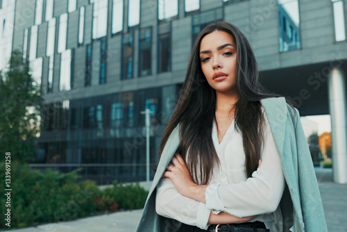 Smiling girl with brown hair stands of corporate building for break dressed in smart shirt and jacket thrown over back, walks outside past glass skyscraper, secretary, businesswoman, boss