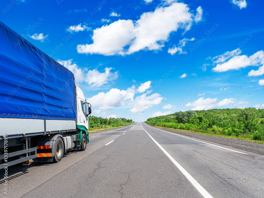 A large cargo truck with a blue tarp-covered trailer travels on a straight highway, surrounded by lush green scenery under a bright blue sky with scattered clouds