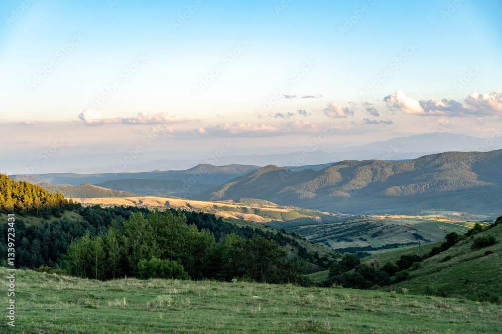 Fototapeta premium Rolling Hills and Majestic Mountains: A Scenic Escape Experience tranquility with this vibrant image showcasing lush green hills, scattered trees, and shrubs in the foreground. Towering hills and maje
