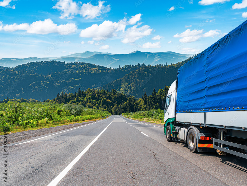 A large truck with a blue tarp-covered cargo area drives along a scenic highway, surrounded by lush green hills and mountains under a bright blue sky.