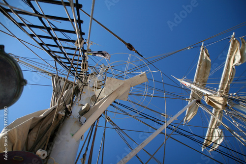 Une vue en contre-plongée du mât d'un voilier mettant en valeur les cordages, poulies et voiles partiellement hissées sous un ciel bleu éclatant.