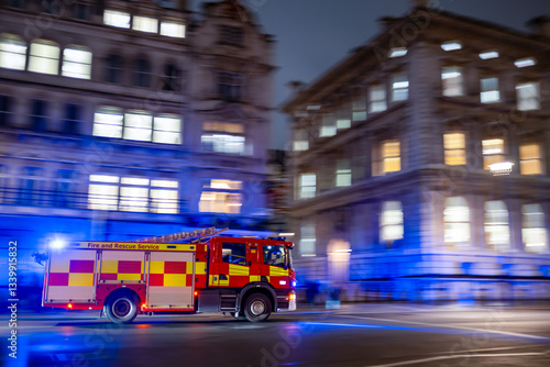 Fast red firetruck moving on emergency call in London city center with tall bouldings around. lights on and firefighters inside. motion effect. panning effect. fire brigade UK	
