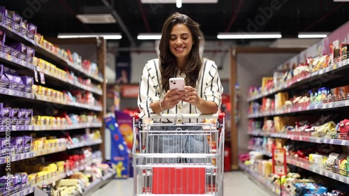 Happy young woman texting on smartphone while shopping at the supermarket