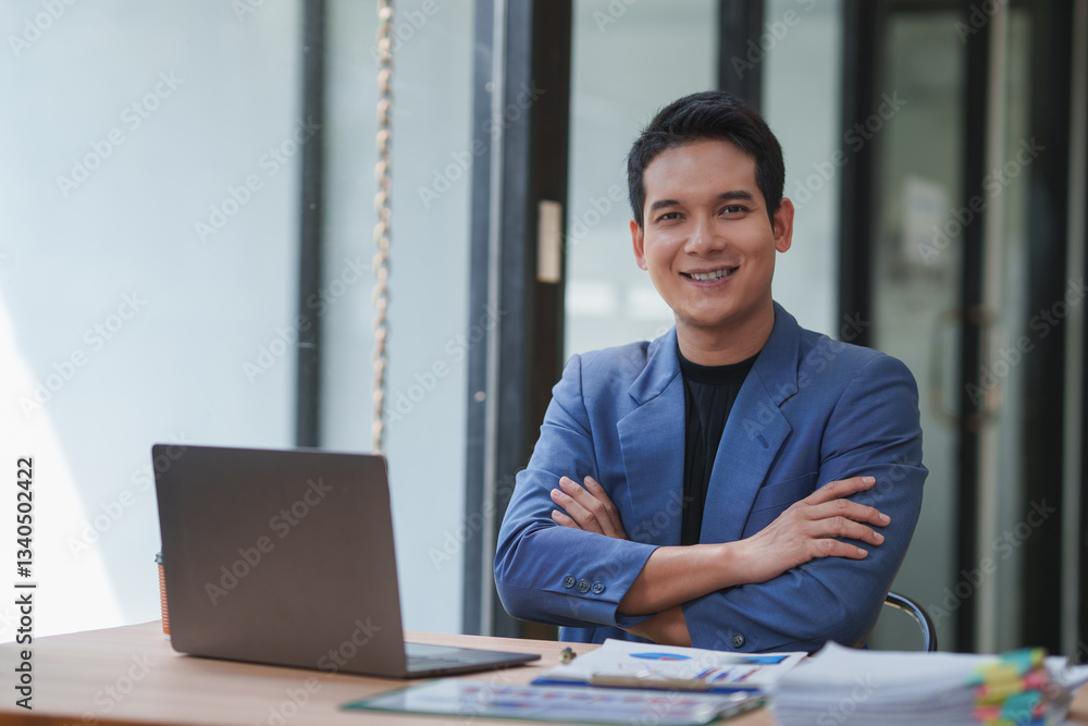 Fototapeta premium Confident young entrepreneur smiling while sitting at his desk in a modern office, with arms crossed and laptop and paperwork in front of him, showcasing professionalism and success in business