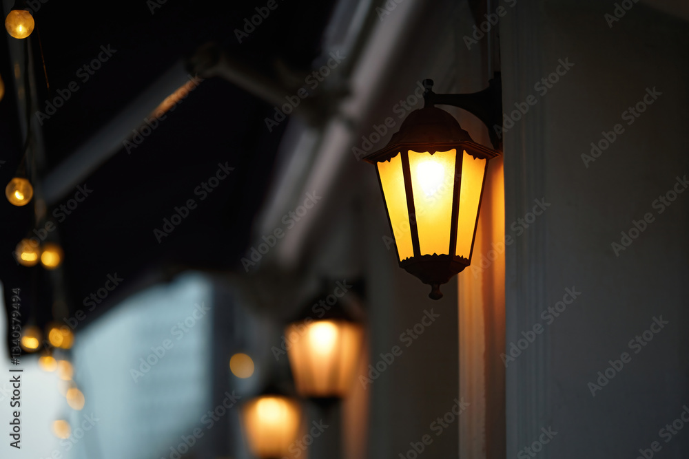 Vintage lantern glowing warmly on building exterior at night, Cozy, nostalgic atmosphere with soft ambient lighting and classic architectural charm. Illuminated wall-mounted lantern. Selective focus