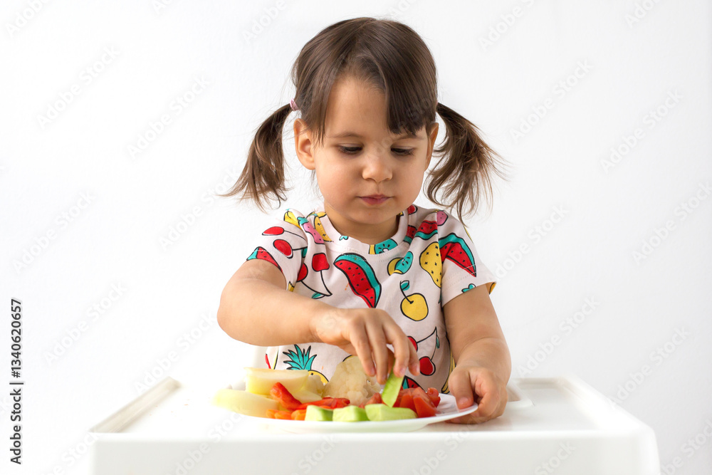 Little girl with pigtails sitting in high chair, carefully picking up piece of cucumber from plate of fresh vegetables. Healthy eating, childhood nutrition, independent eating Little girl with pigtails sitting in high chair, carefully picking up piece of cucumber from plate of fresh vegetables. Healthy eating, childhood nutrition, independent eating