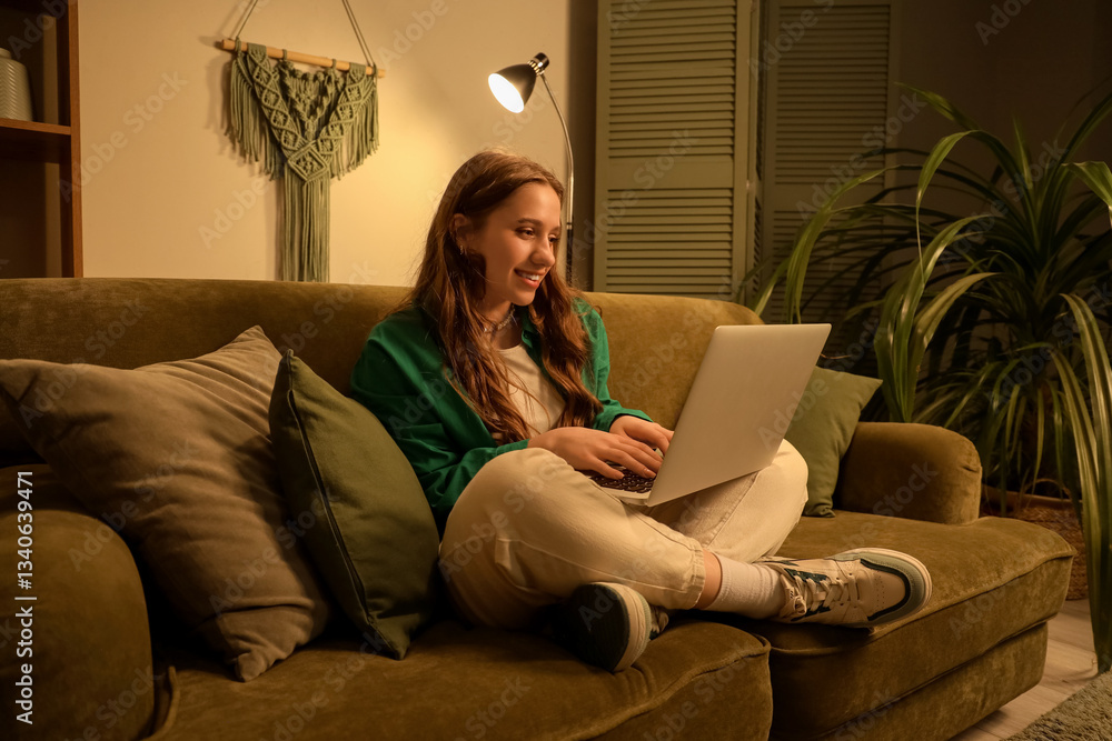 Fototapeta premium Young woman using laptop on sofa at home in evening