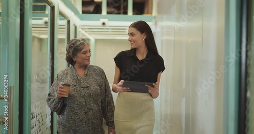 Indian older woman lady talk gossip young adult staff discuss work day time task indoor office place smiling female hold paper cup drink coffee expert girl hold tablet device walk at hall way space