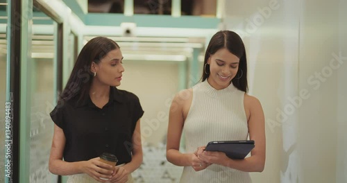 Indian asian happy young adult gen z two girl woman lady staff use tablet device talk discuss task job office work place smiling female drink coffee cup while walking fun joy indoor space break time