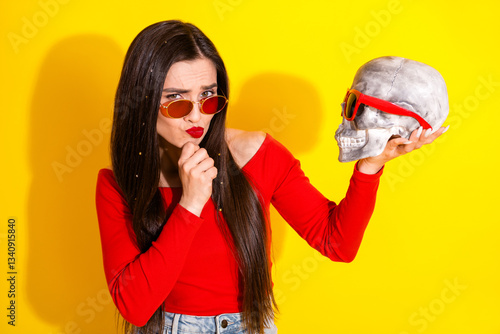 Fashionable young woman holding a sculptural skull wearing sunglasses against a vibrant yellow background