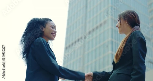  Professional businesswomen in formal attire shaking hands in front of a modern skyscraper, symbolizing a successful deal, collaboration, or networking. 
