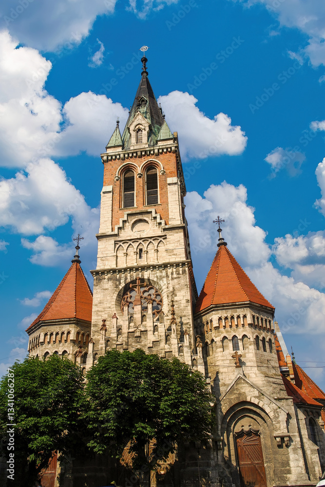 Fototapeta premium Majestic Saint Stanislaus Church with ornate tower and red-roofed turrets against a blue sky with white clouds in Chortkiv, Ternopil region, Ukraine. Gothic Revival architecture