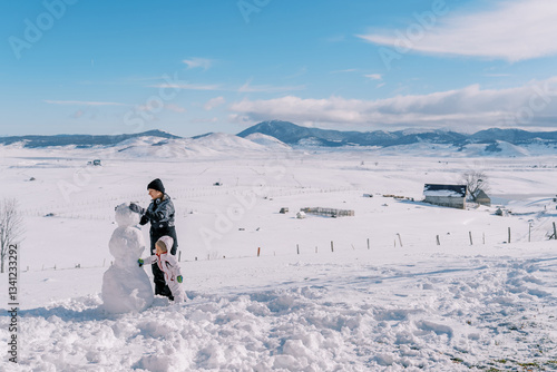 Little girl helps her mother build a snowman in a mountain valley. Side view