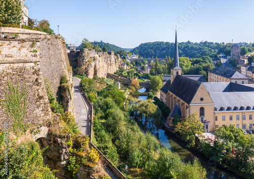 Panorama over the Alzette valley below the old town in Luxembourg City, Luxembourg, with the Neimënster Abbey, the Bock rock (left) and the Jacob Tower (right) in the distance on a sunny day.