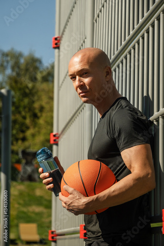 Obraz na płótnie Portrait of basketball player or trainer in black t-shirt holding orange basketball ball and water bottle on sports field. Outdoors. Basketball coach. w salonie