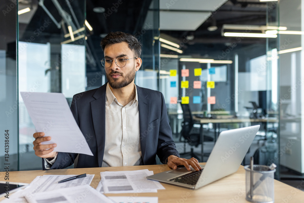 A thoughtful businessman reviews documents and uses his laptop, working in a modern office setting with a glass-walled conference room in the background A thoughtful businessman reviews documents and uses his laptop, working in a modern office setting with a glass-walled conference room in the background
