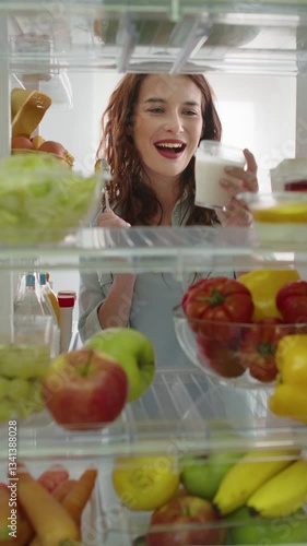 Beautiful young woman opens fridge door, looks inside takes out a yogurt, preparing healthy meal using fresh groceries. Point of View POV shot from inside a kitchen refrigerator full of healthy food