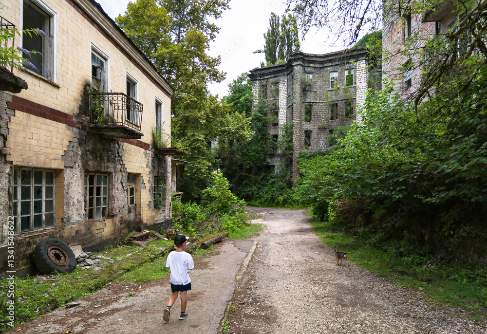 Child kid walks on abandoned ruined street in Akarmara town, Tkvarcheli city, Abkhazia. Old building overgrown by green grass in summer. Concept of war