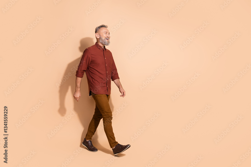 Fototapeta premium Middle-aged man in casual attire walking confidently against a beige background, showcasing modern fashion and leisure.