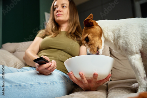 Woman relaxing on sofa with remote control while dog steals food. Pet dog eat food from bowl while owner focuses on TV. Home life and pet behavior concept