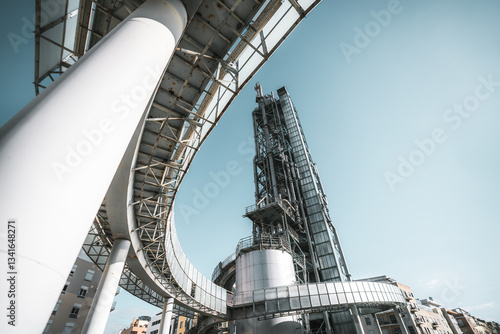 Low-angle view of modern industrial architecture featuring steel beams, curved elevated walkway, futuristic metallic structures, and buildings against clear blue sky background; wide-angle shot