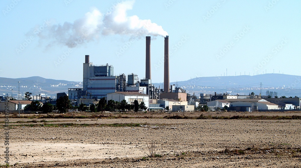 polluted industrial area with smokestacks emitting thick smoke into the air, surrounded by barren land and a nearby residential neighborhood [Carcinogens]:[Impact on population quality of life] 