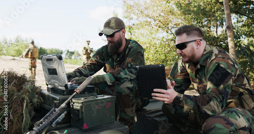 A powerful military unit at the training ground, dressed in camouflage uniforms, helmets, and backpacks, holds a tablet, enters tactical data, and discusses strategy to fight and surprise the enemy.