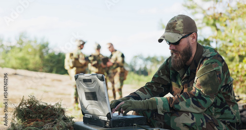 Armed soldiers, prepared for battle, stand in formation in the field, waiting for orders as the commander at the computer updates and transmits data to the base.