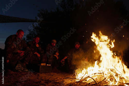 A large bonfire flame, behind it military men in camouflage uniforms are sitting, talking and warming themselves at night by the hearth