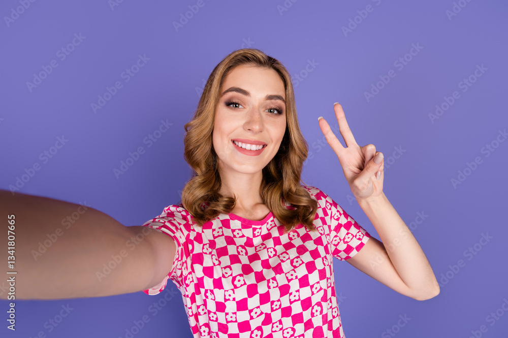 Fototapeta premium Smiling young woman taking selfie with peace gesture wearing pink checkered shirt in front of violet studio background