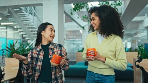 Two women gesturing while having warm conversation. Walking together in middle of office while holding cups with warm drinks. Looking at each other and smiling. Enjoying morning at work.