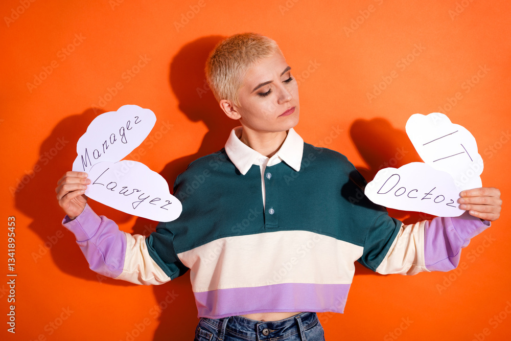 Young woman holding career path options on paper cutouts, standing against orange background with casual fashion expression Young woman holding career path options on paper cutouts, standing against orange background with casual fashion expression
