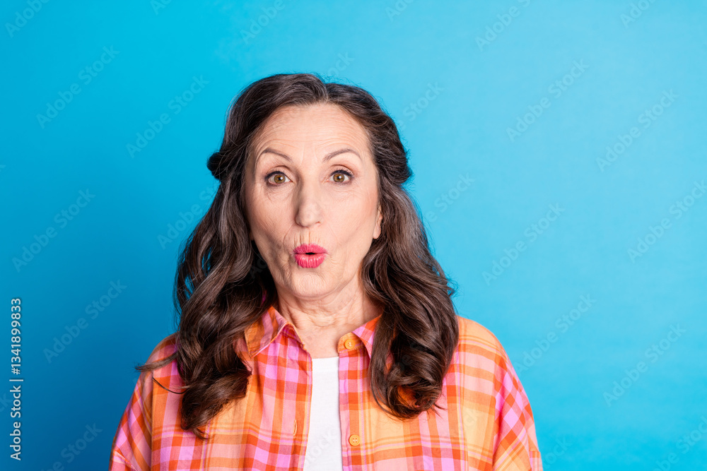 Fototapeta premium Mature woman expressing surprise on a blue background while wearing a colorful checkered shirt