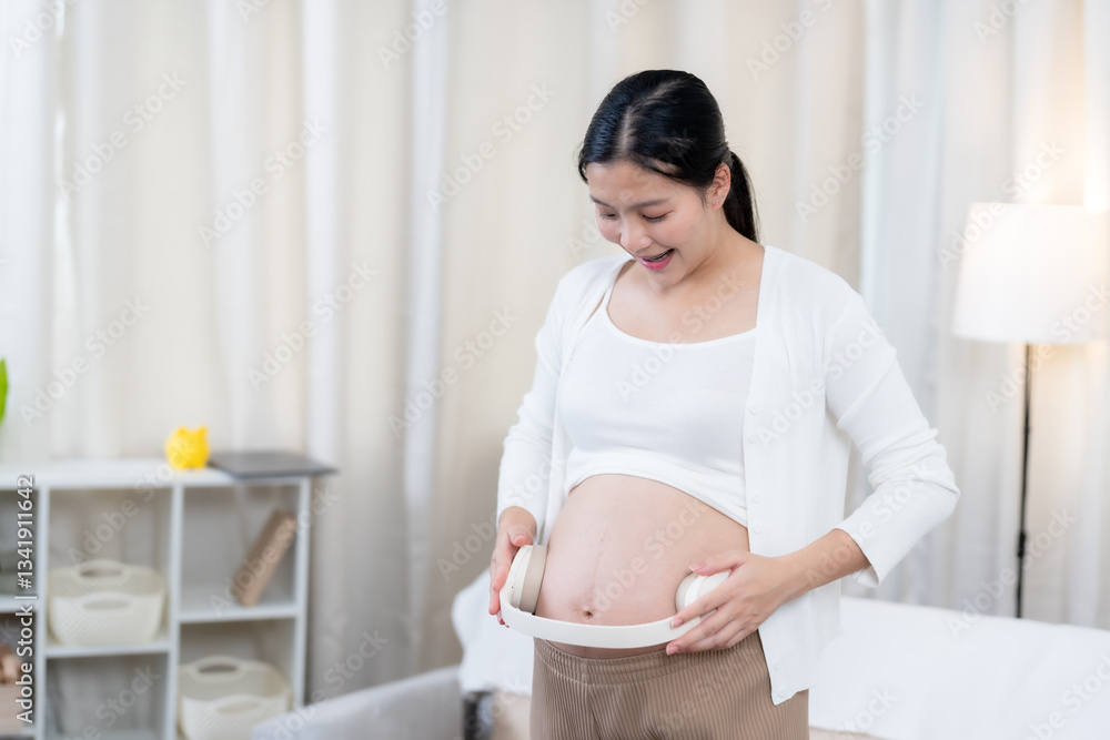 Asian pregnant woman placing wireless headphones on belly, playing music for baby with a warm smile. Wearing a white cardigan and casual outfit in a comfortable and serene home environment. Asian pregnant woman placing wireless headphones on belly, playing music for baby with a warm smile. Wearing a white cardigan and casual outfit in a comfortable and serene home environment.