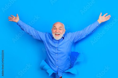 Happy old bearded man with blue contact lenses looking through hole in paper. Smiling senior man in stylish shirt raised hands. Handsome male peeking from ripped paper hole. Season sale and discount. photo