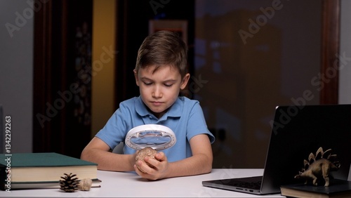 Young boy is sitting at a desk, carefully examining a stone through a magnifying glass. He is focused on his exploration, surrounded by books and a laptop