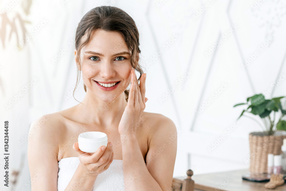 Attractive young woman smiling while applying face cream at home, copy space Attractive young woman smiling while applying face cream at home, copy space