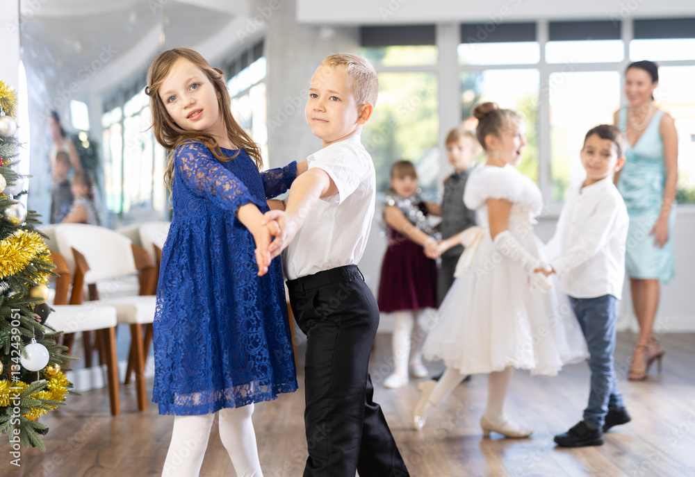 Positive elementary students in festive attire rehearsing ballroom dance number for upcoming Christmas party at school with interested female teacher in background.. Positive elementary students in festive attire rehearsing ballroom dance number for upcoming Christmas party at school with interested female teacher in background..