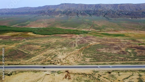Aerial shot of a highway crossing an expansive rural landscape with rolling fields in shades of green and brown. Distant mountains and a reflective water body add depth, creating a tranquil countrysid